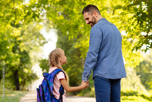 Happy middle aged father and daughter schoolgirl walking to school, holding hands and looking at each other