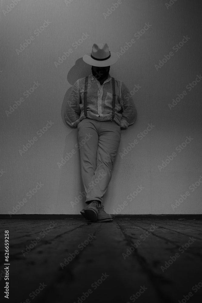 A strict man with a beard in a white suit and white hat stands cross-legged and leans against the wall. The gaze is directed to the floor, hands in pockets. Vertical black and white photo.