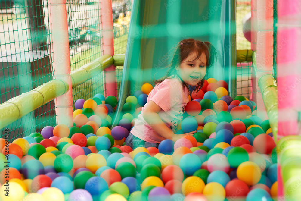 Obraz premium Children's street play attraction fenced with a net. A little girl rolled down the slide and is floundering among many colorful balls.