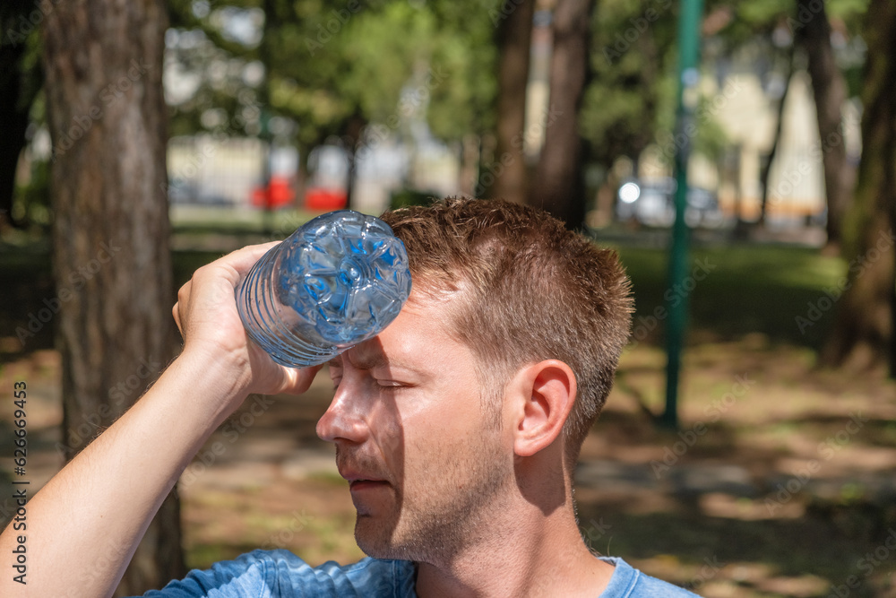 Man suffering from heat wave Charon in South Europe. Caucasian male presses bottle of water to face to cool off suffering from heat sitting outdoors - obrazy, fototapety, plakaty 