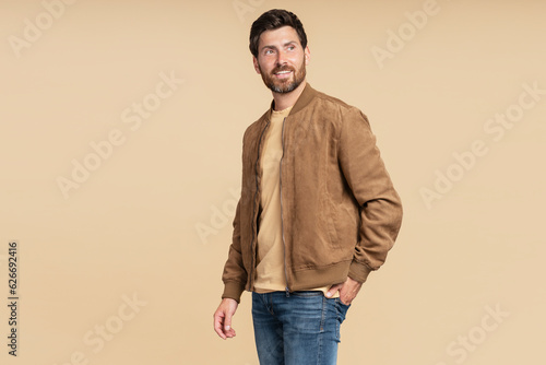 Handsome smiling bearded man wearing brown autumn jacket, stylish jeans isolated on beige background. Portrait of successful middle aged fashion model posing for pictures, studio shot