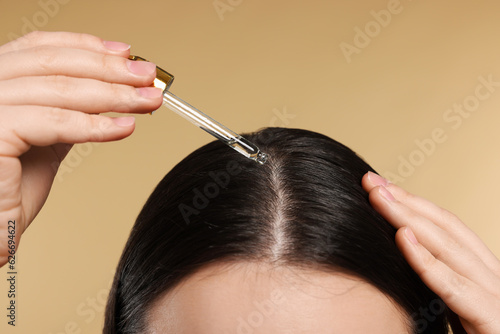 Woman applying essential oil onto hair roots on beige background, closeup