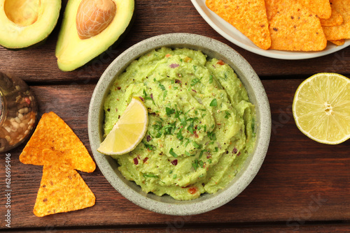 Bowl of delicious guacamole, lime and nachos chips on wooden table, flat lay