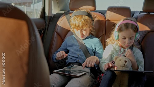 Preteen siblings at back seat of car using digital tablet for watching movies, listening to the music in headphones, surfing social media, e-learning online from home. Concept of happy childhood.