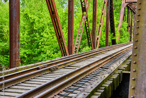 Wall of green leaves behind rusty iron beams of truss railroad bridge with detailed train tracks