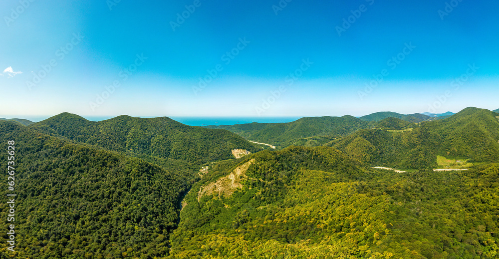 Panorama aerial view of the gorge of the mountain river Shepsi surrounded by the mountains of the Caucasus covered with green forest and the blue sea in the distance on a sunny summer day with a blue 