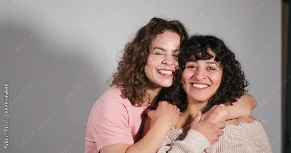 A happy young Latina/multiracial lesbian couple smiles, hugs, and snuggles up next to each other in a studio. A happy young Latina/multiracial lesbian couple smiles, hugs, and snuggles up next to each other in a studio.