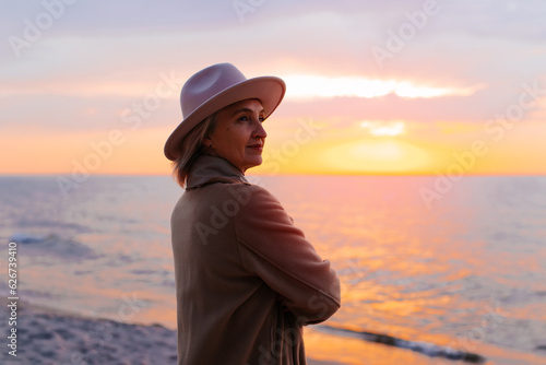 Senior stylish woman clasping her hands together standing by sea at sunset and enjoying evening looking away