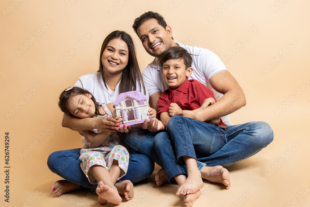 Happy young indian parents and kids wearing casual cloths holding house sitting together on floor isolated over beige studio background. Asian Family bonding. Concept of real estate and house buy.