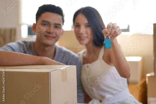 Happy asian couple holding key of new house. Wife and husband moving home, packing cardboard boxes together. Homeowners celebrating new purchase buying real estate.