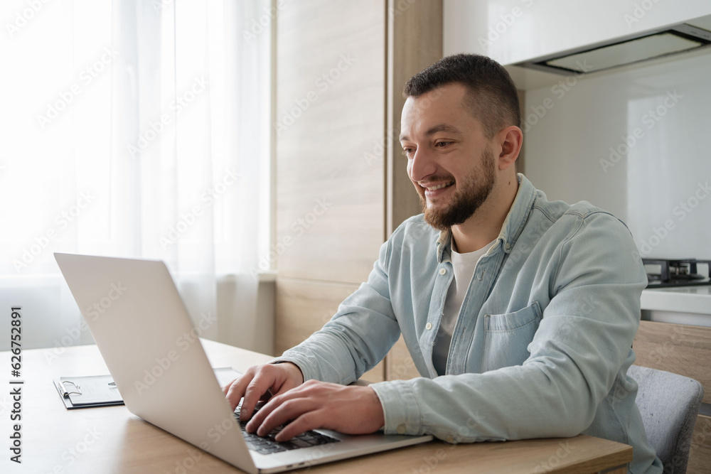 Portrait of young smiling man using laptop sitting at desk, writing in notebook. Cheerful guy browsing internet, watching webinar studying online, looking at pc screen at home.