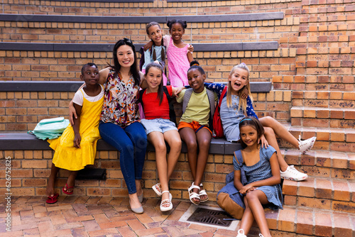Portrait of diverse female teacher and schoolgirls sitting in elementary school outdoor auditorium