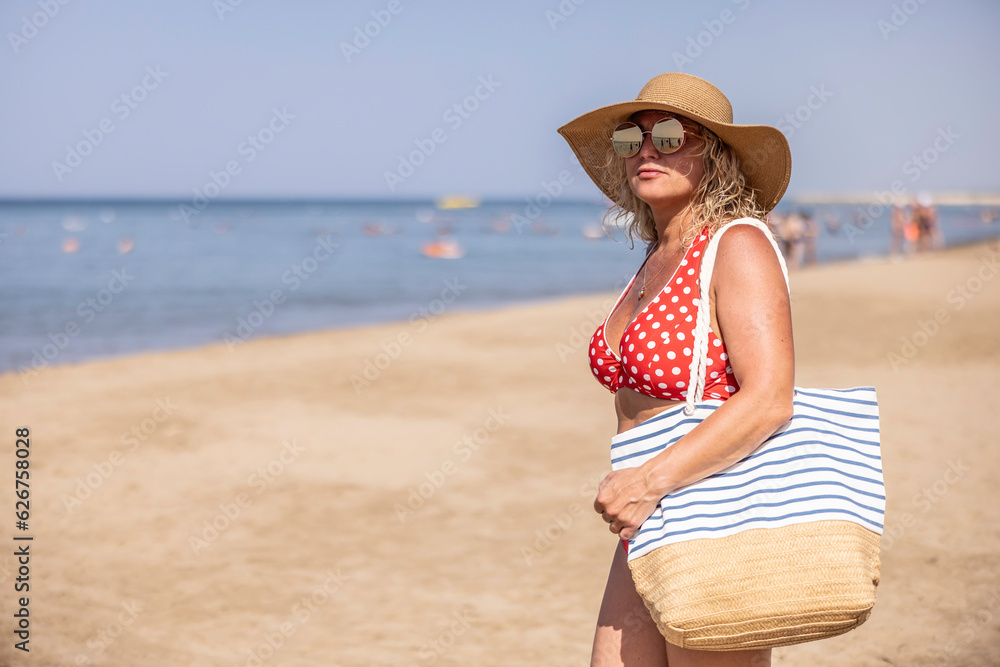 Good looking middle aged woman with beach bag in retro swimsuit wearing hat and sunglasses on the beach by the sea.