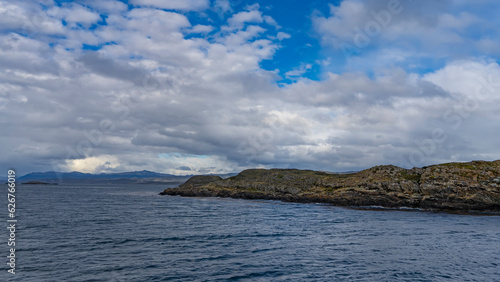 A rocky island in the ocean is covered with sparse stunted vegetation. Ripples on the water. Clouds in the blue sky. The Beagle Channel. Argentina.