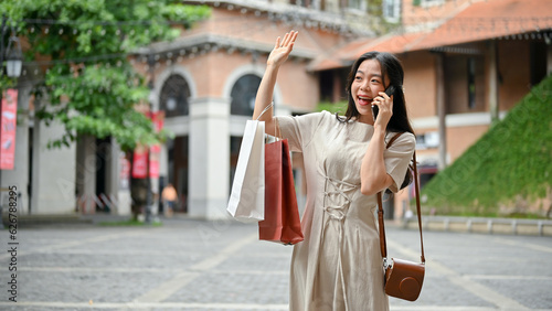 A happy Asian woman with her shopping bags is talking on the phone with her friend on the street.