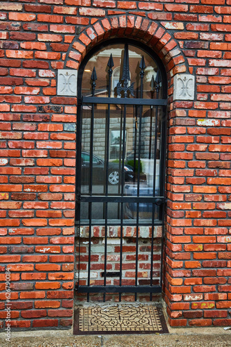 Red brick wall with black metal gate blocking doorway now bricked up with window and metal floormat