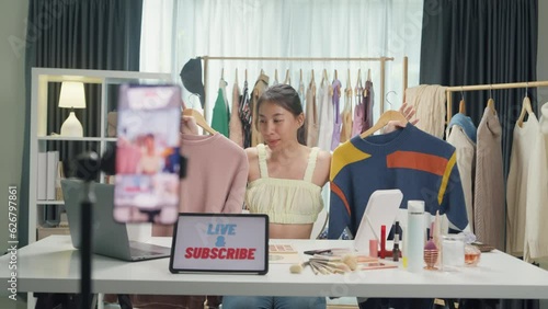 Young Asia lady seller look at phone camera live stream use laptop answer follower comment introduce sweater new collection new arrival on her chanel at studio. Woman fashion online shop concept.