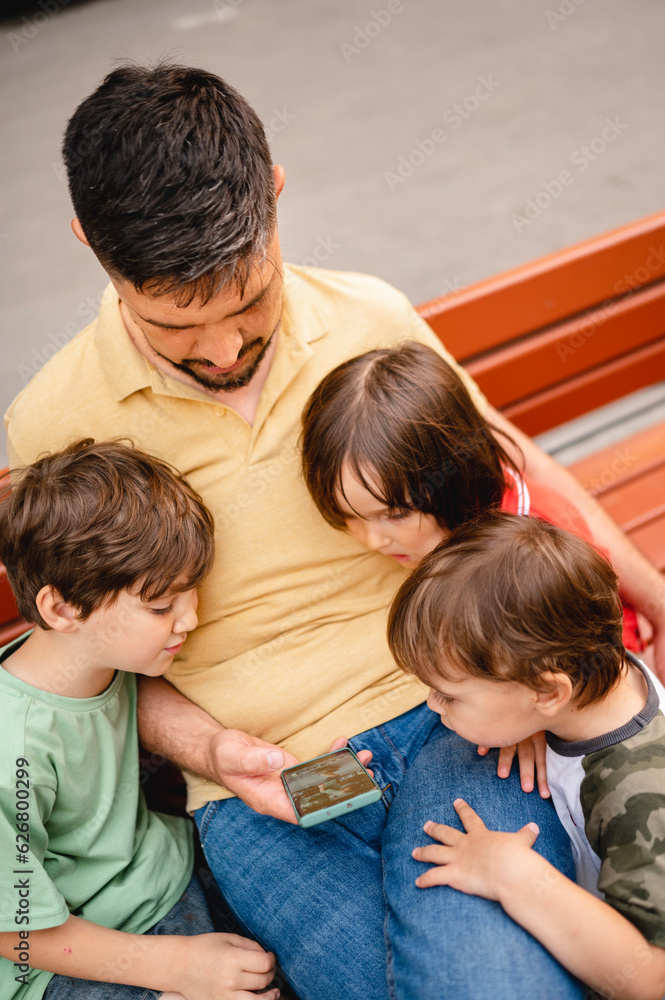 Father sharing smart phone with sons sitting on bench at park Father sharing smart phone with sons sitting on bench at park