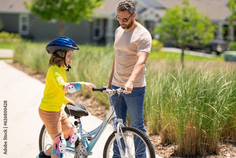 Fototapeta premium Father and son in bike helmet for learning to ride bicycle at park. Father helping son cycling. Father and son on the bicycle on summer day. Little son trying to ride bike with father. Fathers day.