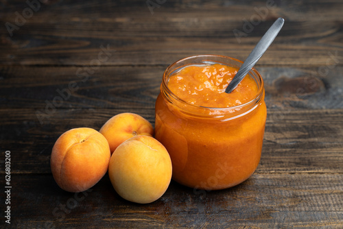 Apricot jam in a glass jar with a spoon inside, fresh apricots on a wooden background