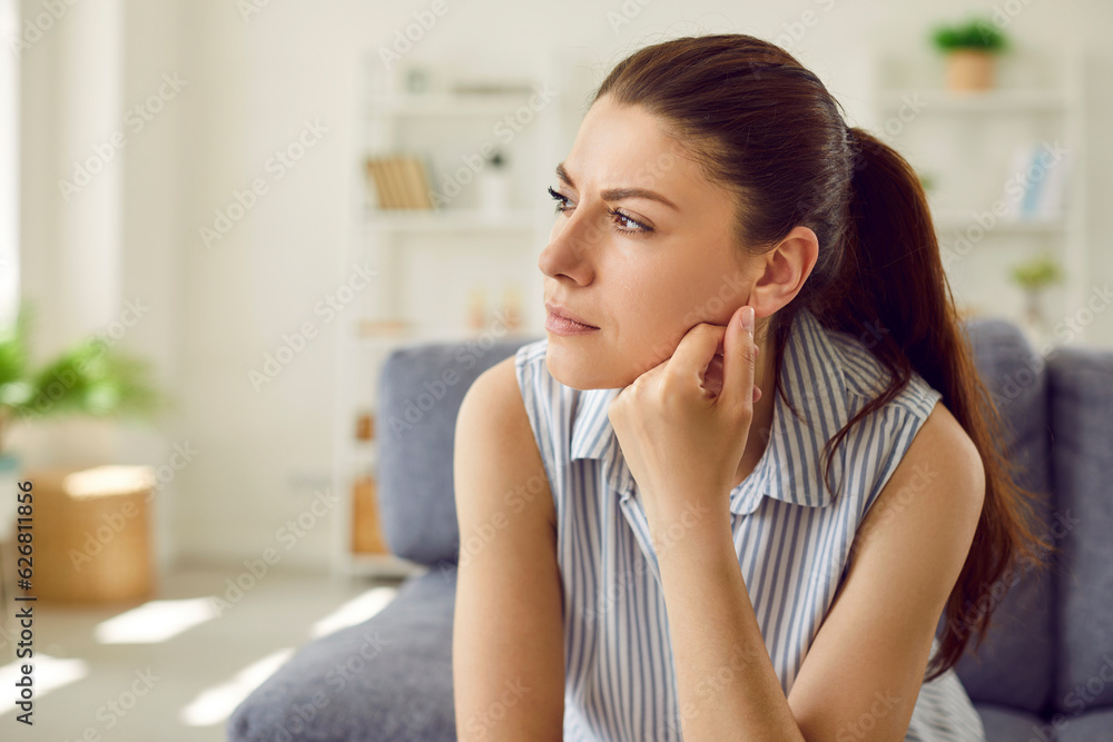 Close-up portrait of upset sad depressed woman sitting on the sofa at home in bad thoughts alone. Thoughtful young girl feel lonely indoors with problems in relationships or dreaming about something.