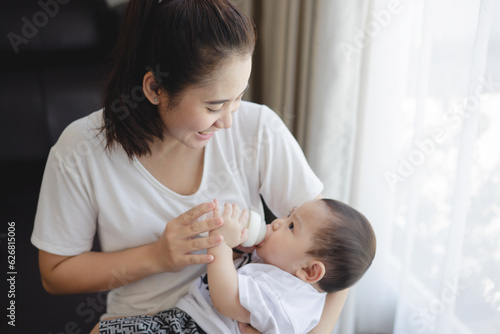 Asian young mother holding and feeding baby from milk bottle at living home. loving woman giving drink milk to her son and she feels happiness.
