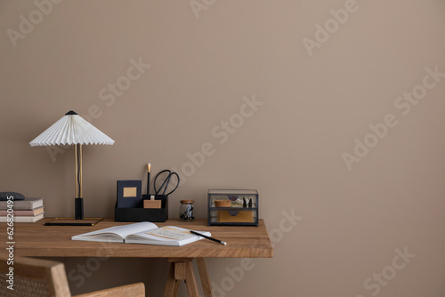 Fotografia Minimalist composition of warm workspace interior with copy space, wooden desk, rattan armchair, stylish lamp, books, glass casket, brown wall and personal accessories