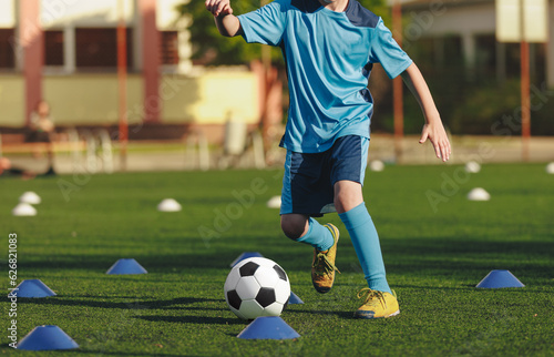 Physical education training class for children. Kids kicking classic soccer ball on slalom drill. Legs of school boy running fast and kicking soccer ball between red training cones