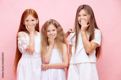 Portrait of cheerful little girls, kids covering mouth with hands, hiding smile and laugh against pink studio background. Concept of skincare, childhood, cosmetology, health, beauty, wellness, ad