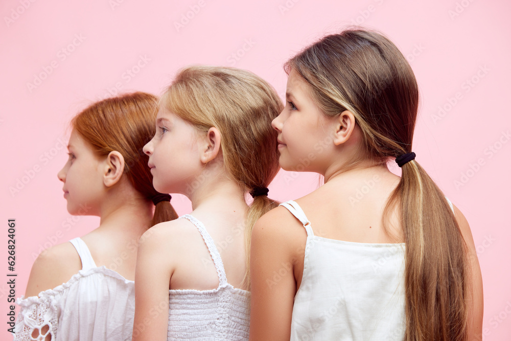Back view portrait of beautiful three little girls, children in white clothes, with ponytail posing against pink studio background. Concept of skincare, childhood, cosmetology and health, beauty, ad Back view portrait of beautiful three little girls, children in white clothes, with ponytail posing against pink studio background. Concept of skincare, childhood, cosmetology and health, beauty, ad