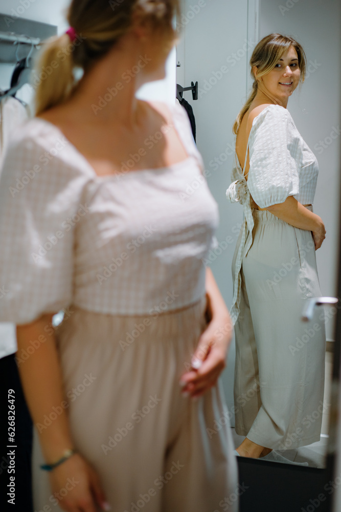 Fototapeta premium Adorable woman in new white blouse and being trousers smiling while checking outfit in reflection in dressing room.