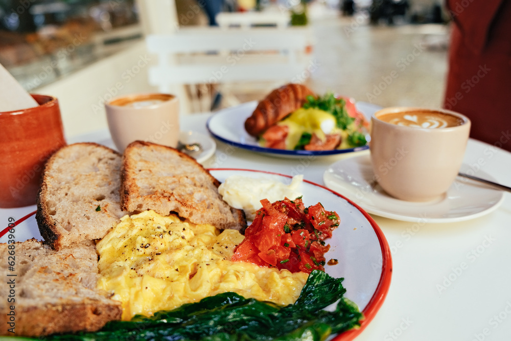 Brunch in an outdoor cafe - bread, scrambled eggs, tomatoes, butter, croissant, salmon, eggs benedict, coffee on a white table