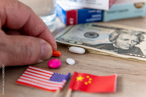Photography A pharmaceutical, medical and drugs conflict concept with the American and Chinese flags, US Dollar bills and various medicines on a desk with a person taking a tablet