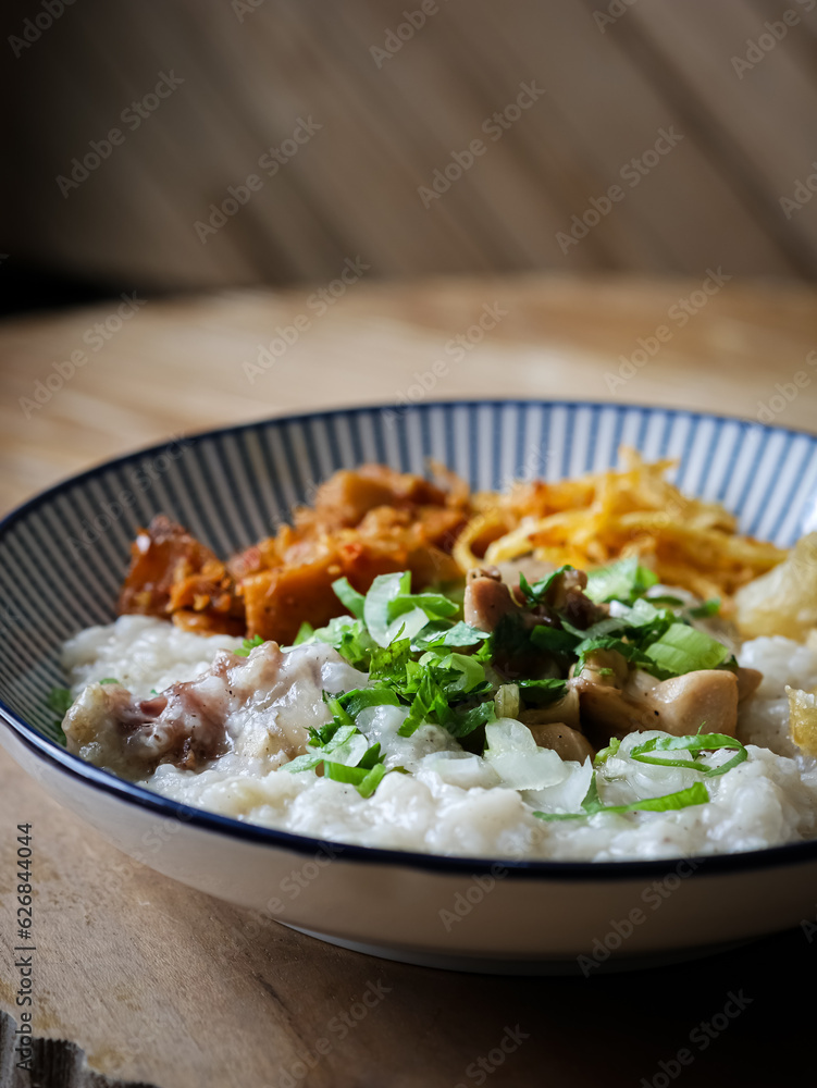 Bubur ayam or chicken porridge is Chinese Indonesian rice porridge topped with various savoury condiments served on white bowl and wooden table. Bubur ayam or chicken porridge is Chinese Indonesian rice porridge topped with various savoury condiments served on white bowl and wooden table.