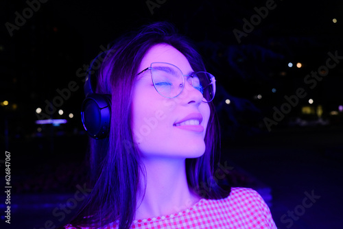Young and happy woman listening to music on headphones at night on the street and smiles