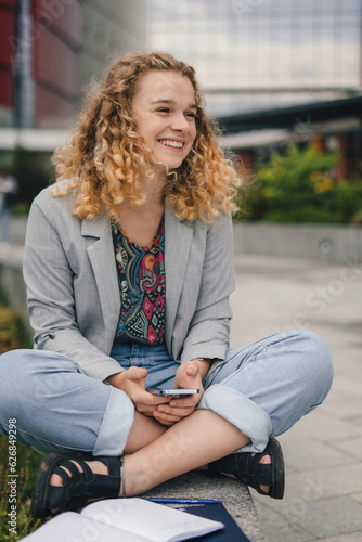 Cheerful smiling woman wearing jacket using mobile phone outside the campus looking away. Technology concept.