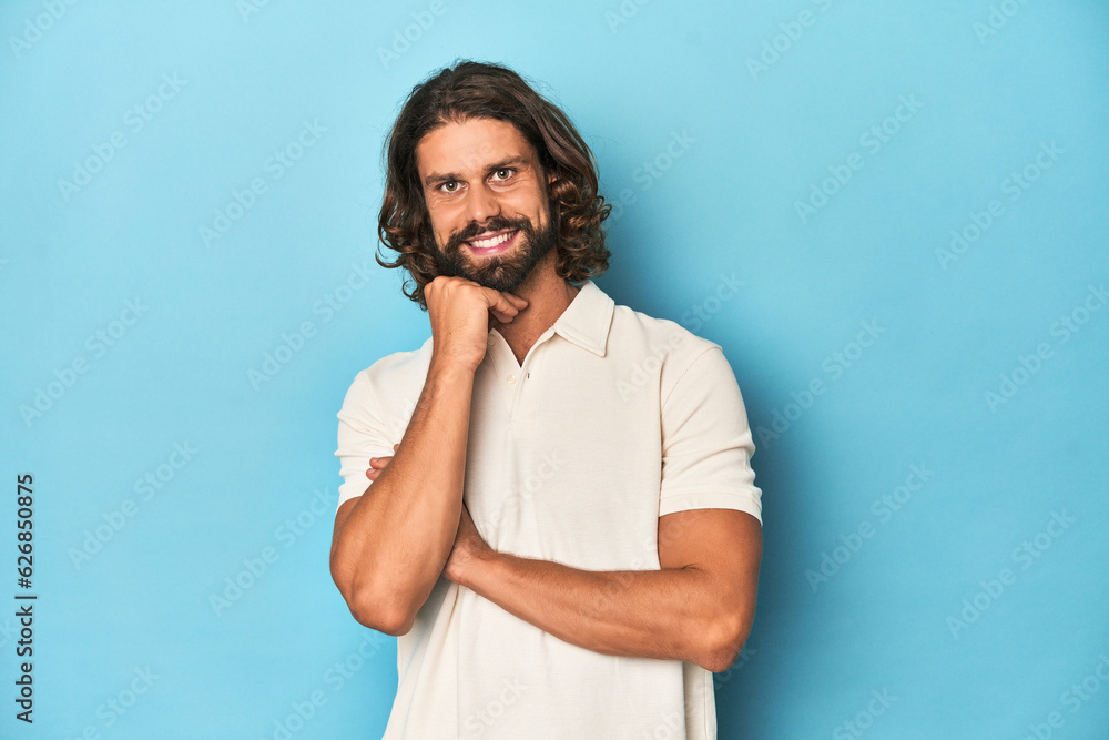 Long-haired man in a white polo, blue studio smiling happy and confident, touching chin with hand. Long-haired man in a white polo, blue studio smiling happy and confident, touching chin with hand.