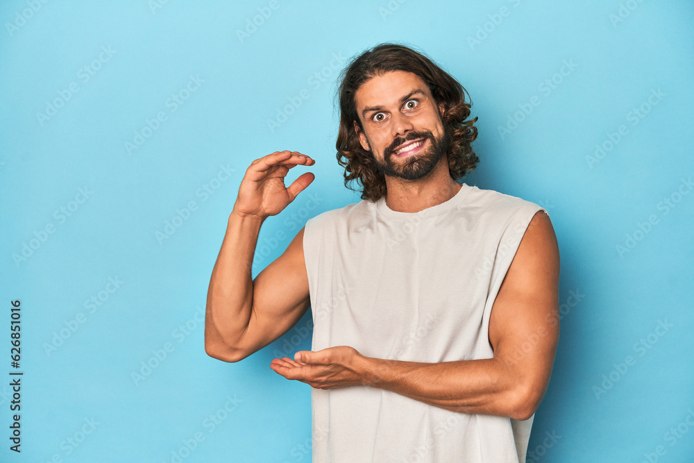 Bearded man in a tank top, blue backdrop shocked and amazed holding a copy space between hands. Bearded man in a tank top, blue backdrop shocked and amazed holding a copy space between hands.