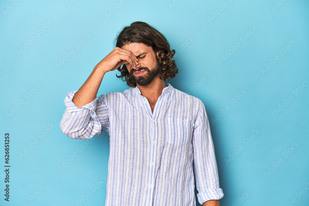 Man with beard in blue striped shirt, blue studio having a head ache, touching front of the face. Man with beard in blue striped shirt, blue studio having a head ache, touching front of the face.