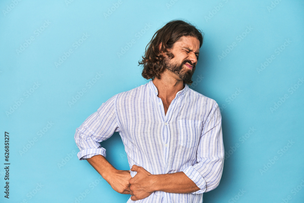 Man with beard in blue striped shirt, blue studio having a liver pain, stomach ache. Man with beard in blue striped shirt, blue studio having a liver pain, stomach ache.