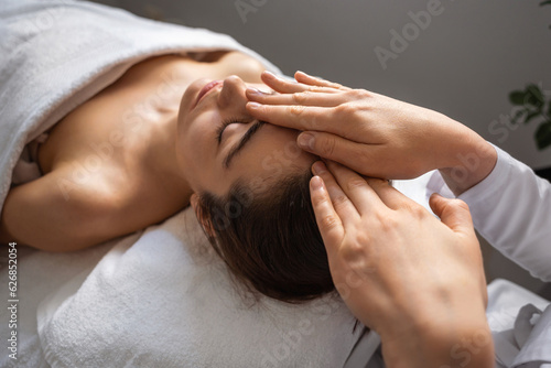 Hands of female masseuse doing a massage on the forehead of a beautiful young woman lying on a massage table with closed eyes in a spa salon. Beautiful young