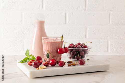 A freshly made homemade smoothie made with ripe cherries in a large glass glass with almonds and mint and a full bottle of smoothie. White brick wall