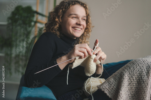 Curly haired woman sitting on the sofa in the living room and knitting a scarf for the winter. Hobby for young woman.
