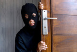 The robber man wearing mask and black clothes hiding behind the door and looking at camera