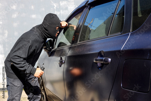 Man dressed in black wearing mask hold screwdriver, looking through car window and wondering how to break into the car. Car thief, car theft concept