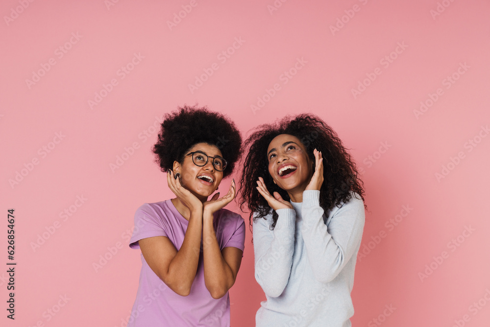 Two smiling african women looking up and holding hands at their faces while standing isolated over pink background Two smiling african women looking up and holding hands at their faces while standing isolated over pink background