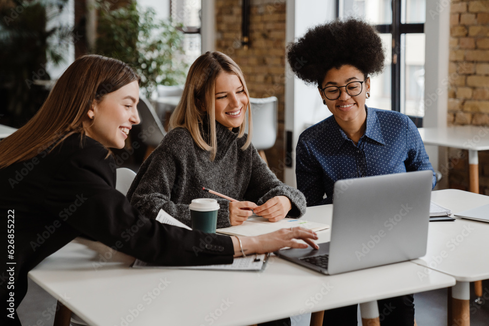 Three smiling women using laptop while working together in office Three smiling women using laptop while working together in office