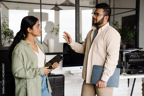 Two young colleagues having business conversation while standing in office