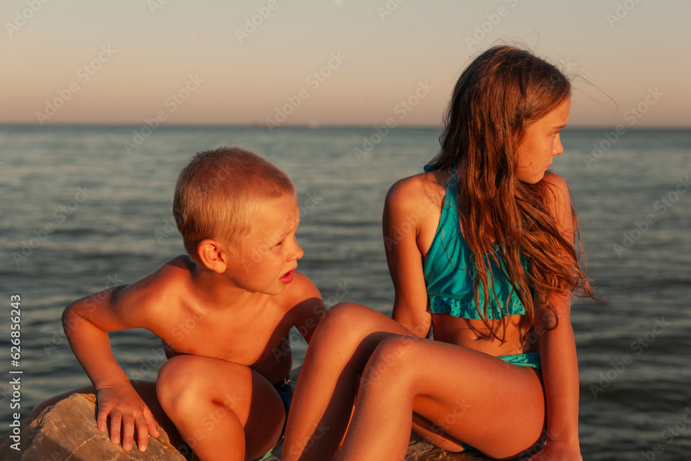 Brother and sister are sitting on the rocks on the seashore, at sunset. Girl with long hair, blond boy. Both looking at the sunset Brother and sister are sitting on the rocks on the seashore, at sunset. Girl with long hair, blond boy. Both looking at the sunset