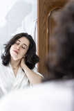 portrait of a young latin woman with a gesture of pain looking at herself in the mirror with her hands on her neck showing neck pain